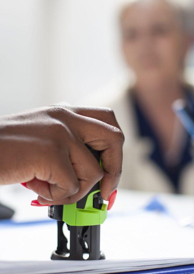 Selective focus on hand of black healthcare specialist applying stamp on document as senior woman watches attentively. Elderly patient waits as african american doctor completes her medical documents.