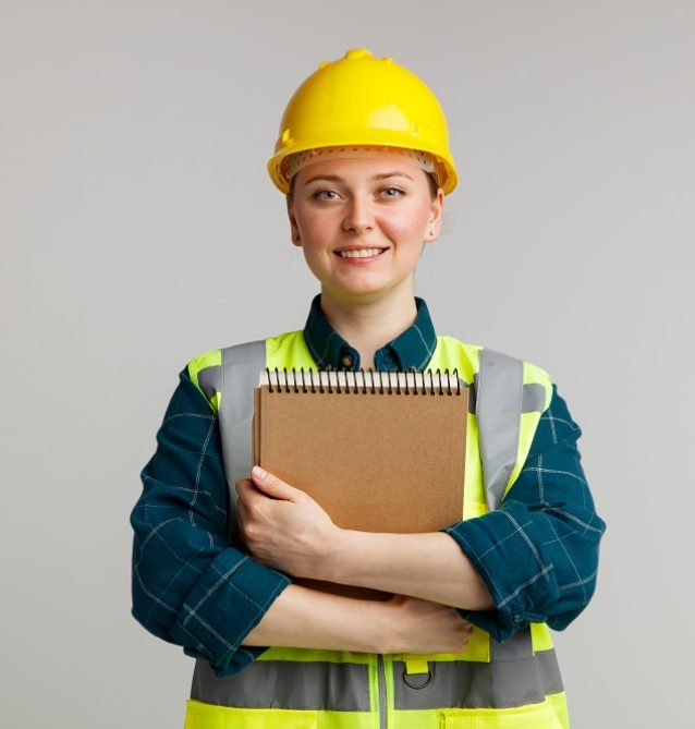 smiling young female construction worker wearing safety helmet and safety vest hugging notepad looking at camera isolated on white background